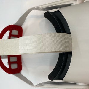 May include: White virtual reality headset with a white elastic strap and a red padded strap. The headset has black foam padding around the eyes.