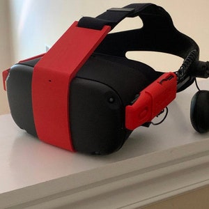 May include: A black virtual reality headset with a red strap and red side grips. The headset has a black strap and a black headband.