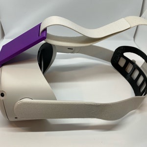 May include: A white virtual reality headset with a purple strap attachment. The headset has a white strap and a black adjustable strap.