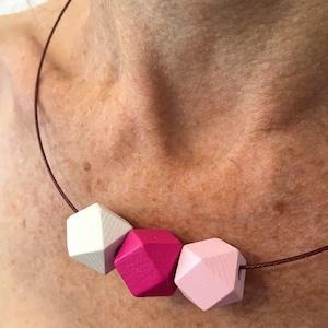 Puede incluir: Un collar con tres cuentas de madera en tonos rosa y blanco. Las cuentas son de forma geométrica y tienen un acabado mate.