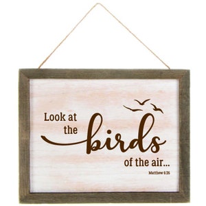 May include: A wooden sign with a white background and a brown wood frame. The sign has a brown inscription that reads "Look at the birds of the air... Matthew 6:26". The inscription is in a cursive font. There are three stylized birds flying above the inscription.