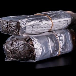 May include: Two packages wrapped in clear plastic, secured with a rubber band. The contents appear dark, possibly granular. One package has a white, perforated material visible. The background is black.