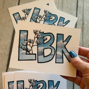 May include: White rectangular cards with the letters "LBK" in a blue watercolor style, filled with a cloudy sky design and cotton branch illustrations. The cards are stacked, with one being held by a hand.
