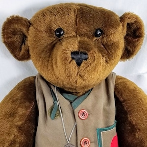 May include: A brown teddy bear wearing a khaki vest with red buttons and a green trim. The bear has black eyes and a black nose. A silver necklace with a pendant hangs from the vest.
