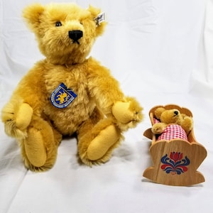 Puede incluir: Un oso de peluche marrón dorado con un botón azul en el pecho se sienta junto a una cuna de madera con un oso de peluche más pequeño dentro. La cuna está decorada con flores rojas y azules.