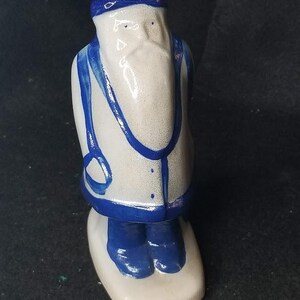 Puede incluir: Figura de cerámica de un marinero, con cuerpo blanco y detalles azules. El marinero lleva un gorro puntiagudo, chaleco con tirantes y botas, todo en azul. La figura se apoya sobre una base blanca.