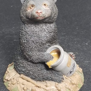 May include: A black bear figurine sitting on a leafy base, holding a honey pot with a gold lid. The bear has a sweet expression on its face and is wearing a brown collar. The base is decorated with leaves and acorns.