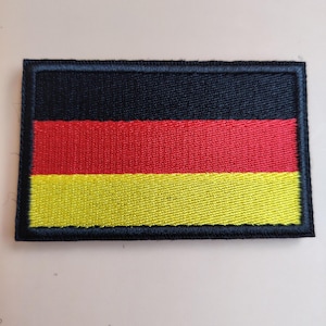 Puede incluir: Un parche rectangular con la bandera alemana. El parche tiene un borde negro y muestra los colores de la bandera: negro en la parte superior, rojo en el medio y amarillo en la parte inferior. El parche es de tela.