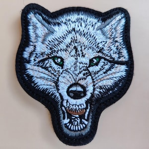 Puede incluir: Parche bordado con la cabeza de un lobo con expresión de gruñido. El lobo tiene pelaje blanco y gris, ojos verdes y un borde negro. El parche está diseñado para ser cosido o planchado sobre tela.