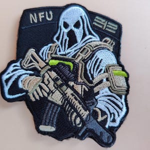 Op de afbeelding: Geborduurde patch met een gestileerde figuur in een witte gehoodde sluier, die een geweer vasthoudt. De patch is zwart met bruine, grijze en groene accenten. De letters "NFU" staan bovenaan. Het ontwerp heeft een militair of tactisch thema.