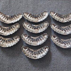 Puede incluir: Diez moldes para hornear de metal de color plateado con forma de media luna. Los moldes son todos del mismo tamaño y tienen una textura ondulada.