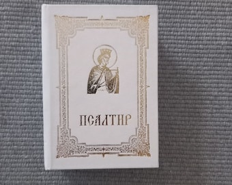 May include: A white book with a gold border and a gold illustration of a person holding a book. The title of the book is "PSALTER" in gold lettering.
