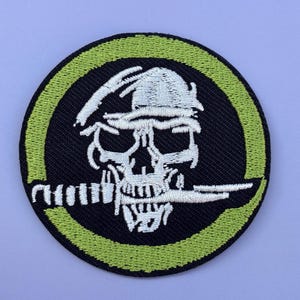 Op de afbeelding: Ronde geborduurde patch met een schedelontwerp. De witte schedel draagt een helm en houdt een mes vast. Het is geplaatst tegen een zwarte achtergrond, met een groene rand. Geschikt voor kleding.