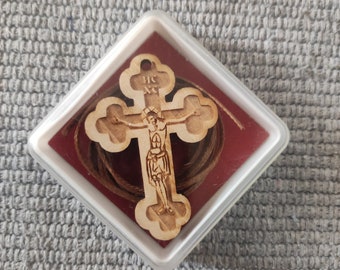 May include: A wooden cross with a carved image of Jesus Christ on it. The cross is in a small, white, square box with a red interior. A brown string is included.