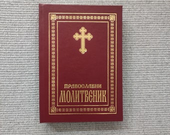 May include: A maroon book with gold accents and text. The cover features a gold cross and decorative border. The text "ПРАВОСЛАВНИ МОЛИТВЕНИК" is printed in gold. The book is rectangular and appears to be a religious text.
