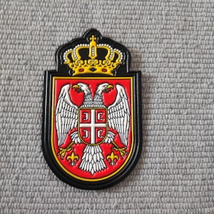 Puede incluir: Un emblema negro, rojo y dorado con un águila bicéfala con alas blancas, una corona y un escudo con una cruz y cuatro Cs. El emblema es un símbolo nacional.