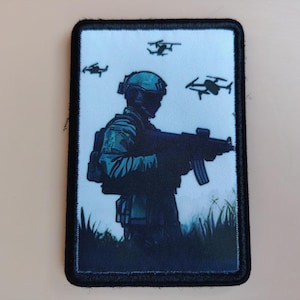 Op de afbeelding: Een rechthoekige patch met een soldaat die een geweer vasthoudt, met drones in de lucht. De soldaat draagt een helm en tactische uitrusting. De achtergrond is lichtblauw met donkerblauwe accenten. De patch heeft een zwarte rand.