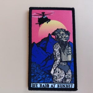 Op de afbeelding: Rechthoekige patch met een zwarte rand met een vrouw in tactische uitrusting met tatoeages, een helikopter en een zonsondergang. De tekst "WE RAID AT SUNSET" staat onderaan. Het kleurenschema omvat roze, blauw en zwart.