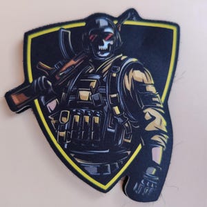 Op de afbeelding: Een zwart en geel embleem met een soldaat met schedelgezicht in tactische uitrusting, die een geweer vasthoudt. Het ontwerp is geplaatst tegen een schildvormige achtergrond met een gele rand. De soldaat draagt een helm, vest en zakken.