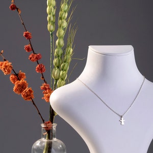 Pode incluir: Um colar de prata com um pequeno pingente de anjo em um busto de manequim branco. Flores secas e hastes de trigo est&atilde;o em um vaso de vidro transparente no fundo.