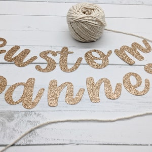 May include: Gold glitter letters spelling "custom banner" on a white wood background with a ball of twine.