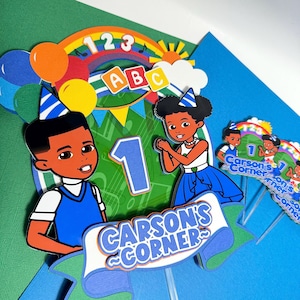 May include: Colorful cake toppers with cartoon illustrations of a boy and girl, a rainbow, balloons, and the number 1. The toppers have the text "Carson's Corner" and ABC letters, perfect for a first birthday celebration.