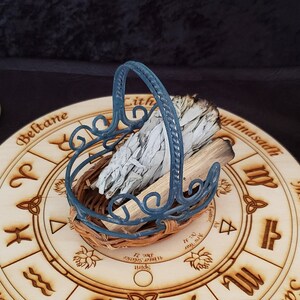 May include: A wooden circle with a wheel of the year printed on it. The wheel of the year is a circular calendar that represents the eight sabbats of the Wiccan year. The circle is decorated with a blue metal basket filled with white sage bundles.