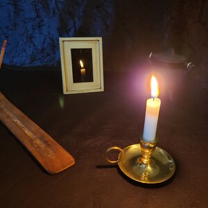 Puede incluir: Una vela encendida en un candelabro de latón se encuentra sobre una superficie marrón oscura, con un palo de incienso de madera y una imagen enmarcada en el fondo.