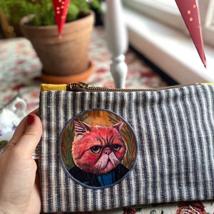 Könnte beinhalten: Eine schwarze und weiße gestreifte Reißverschlusstasche mit einem runden Patch, der eine mürrische orange Katze mit einer blauen Fliege zeigt. Die Katze trägt eine schwarze Jacke und ist von einem goldfarbenen Kranz umgeben.