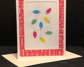 May include: A white card with a red border featuring a cross-stitch design of a string of colorful Christmas lights. The border is decorated with a repeating pattern of small white gift boxes.