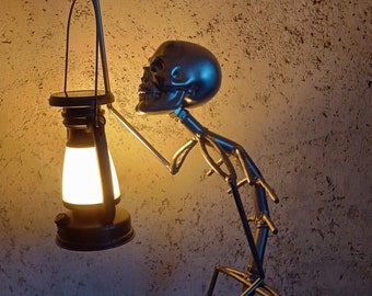 May include: A silver skeleton sculpture holding a black lantern with a white light inside. The skeleton is posed in a standing position with one arm raised and the other holding the lantern. The lantern is lit and casting a warm glow.