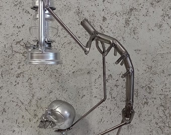 Puede incluir: Una escultura de metal plateado de una persona sosteniendo una linterna y una calavera. La escultura está hecha de varillas de metal soldadas y está de pie sobre una superficie blanca.