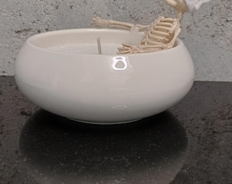 Puede incluir: Una vela en un cuenco de cerámica blanca con una pequeña figura de esqueleto sentada dentro de la cera. El esqueleto está colocado como si se relajara en un baño y lleva una pequeña toalla blanca envuelta alrededor de la cabeza. Se ve una mecha.