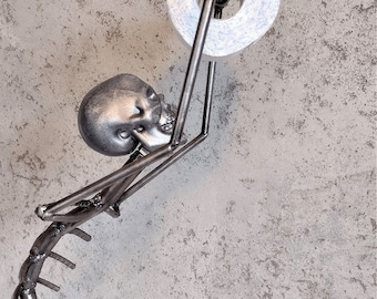 Puede incluir: Una escultura de esqueleto de metal sosteniendo un rollo de papel higiénico. El esqueleto está hecho de varillas de metal soldadas y tiene un cráneo realista. El papel higiénico es blanco con un patrón azul.