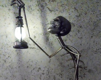 May include: A metal sculpture of a figure holding a lantern with a glowing lightbulb. The figure has a skull-shaped head and is made of welded metal rods. The sculpture is on a white surface against a textured gray background.