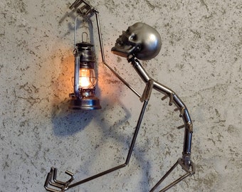Puede incluir: Escultura metálica de una figura con cabeza en forma de calavera, sosteniendo una linterna con una llama encendida. La figura está hecha de varillas metálicas soldadas. La linterna es negra y plateada. El fondo es una pared texturizada gris claro.