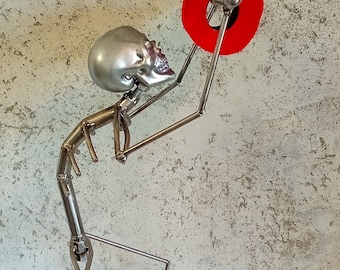 Puede incluir: Una escultura de esqueleto de metal plateado sosteniendo un anillo rojo. La escultura está hecha de finas varillas de metal y tiene una cabeza de cráneo detallada.