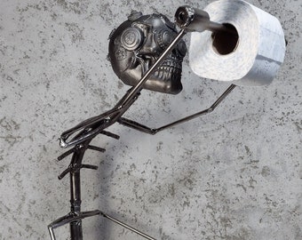 Puede incluir: Una figura de esqueleto de metal sosteniendo un rollo de papel higiénico. El esqueleto está hecho de varillas de metal soldadas y tiene una cabeza de cráneo con engranajes y ruedas dentadas. El papel higiénico es blanco y tiene un núcleo de cartón.