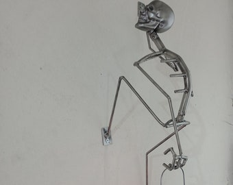 Puede incluir: Una escultura de esqueleto de metal sosteniendo una lámpara de aceite vintage con una llama encendida. El esqueleto está hecho de finas varillas de metal y está fijado a una pared blanca. La lámpara está hecha de metal y vidrio y tiene un globo de vidrio transparente.