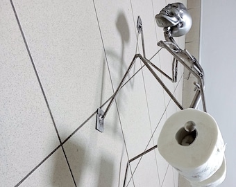 Puede incluir: Un porta rollo de papel higiénico de metal con forma de esqueleto que sostiene un rollo de papel higiénico. El soporte está montado en una pared de azulejos blancos.