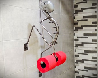 Puede incluir: Una figura de esqueleto de metal que sostiene dos rollos de papel higiénico rojos. El esqueleto está sujeto a una pared con un soporte de metal. El esqueleto tiene una cabeza de calavera plateada.