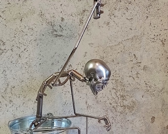 Puede incluir: Una escultura de metal de un esqueleto sosteniendo un rollo de papel higiénico en un cubo. El esqueleto está hecho de metal y está pintado de plata. El rollo de papel higiénico es rojo.
