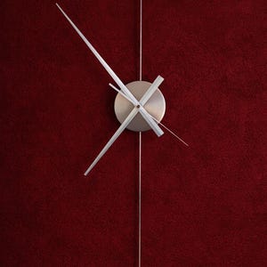 May include: A modern wall clock with a silver face and hands, suspended by a thin silver rod against a deep red textured background. The clock's minimalist design emphasizes its hands and central disc.
