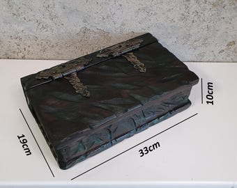 Puede incluir: Una caja oscura y rectangular con una superficie texturizada y bisagras metálicas. La caja mide 33 cm de largo, 19 cm de ancho y 10 cm de alto. La caja probablemente se utiliza para almacenamiento o decoración.