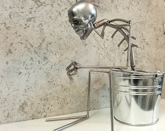 May include: A silver metal skeleton sculpture holding a silver metal bucket. The skeleton is made of wire and has a skull head. The bucket is sitting on a white surface.