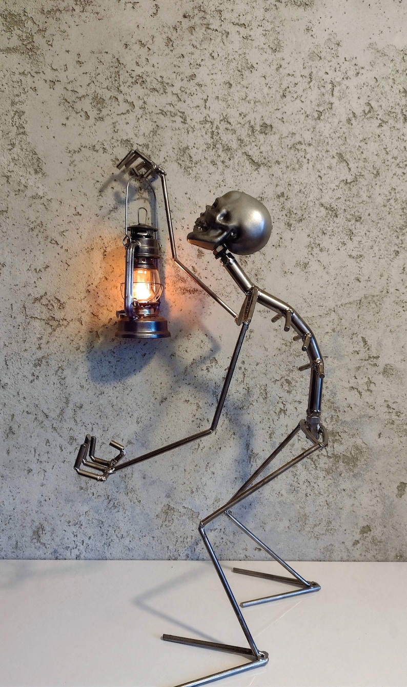 May include: A metal sculpture of a figure holding a lantern with a lit flame. The figure has a skull-shaped head and is made of welded metal rods. The lantern is black and silver. The background is a textured, light gray wall.