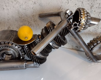Puede incluir: Una escultura de motocicleta hecha completamente de engranajes metálicos y otras piezas industriales. La escultura está pintada de color plateado y tiene un limón amarillo en el asiento.