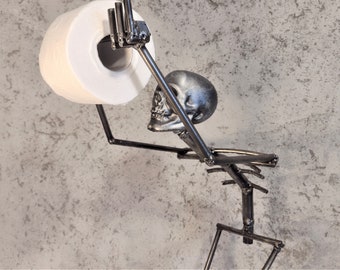 Puede incluir: Un esqueleto de metal sosteniendo un rollo de papel higiénico. El esqueleto está haciendo el dedo medio. El soporte para papel higiénico está montado en una pared.