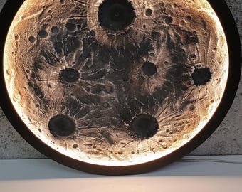 Puede incluir: Una lámpara de pared circular con una superficie texturizada y llena de cráteres que imita la luna. La pieza está encerrada en un marco negro e iluminada desde los bordes, proyectando un brillo cálido que resalta el paisaje lunar oscuro.