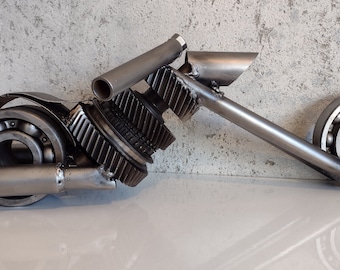 Puede incluir: Una escultura de metal de una motocicleta hecha de engranajes y otras piezas de metal recicladas. La escultura está pintada de color plateado y tiene un diseño realista.